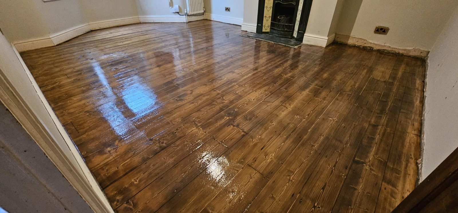 Freshly stained and polished solid pine wood floorboards in a Brockley living room after Wood Floor Sanding, showing a dark, glossy finish around a feature fireplace.